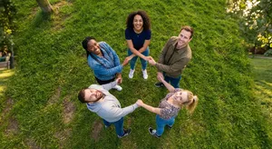 A group of five people joining hands in a circle, seen from above. - Takumi Takumi 1
