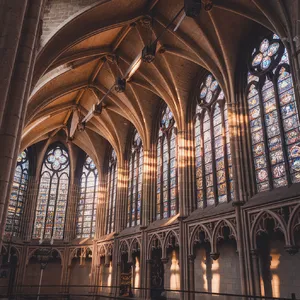 A Gothic cathedral interior at sunset with light streaming through detailed stained glass windows, creating colored patterns on stone floors and columns. Show accurate ribbed vaulting. - Ideogram Ideogram V2